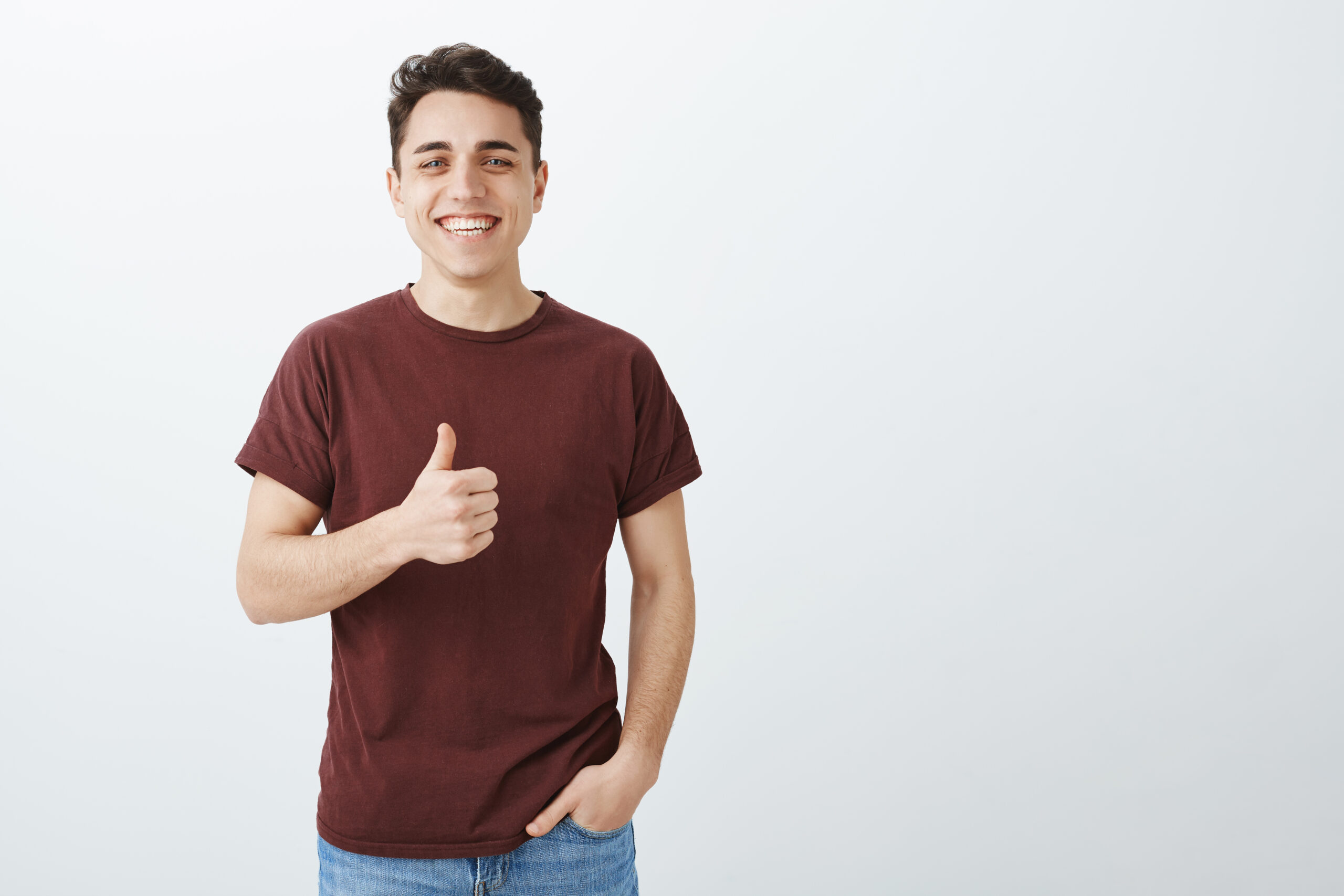 Never heard joke better. Happy handsome man in casual red t-shirt, showing thumbs up and holding hands in pocket, smiling joyfully while liking plan or agreeing with friend over gray background.