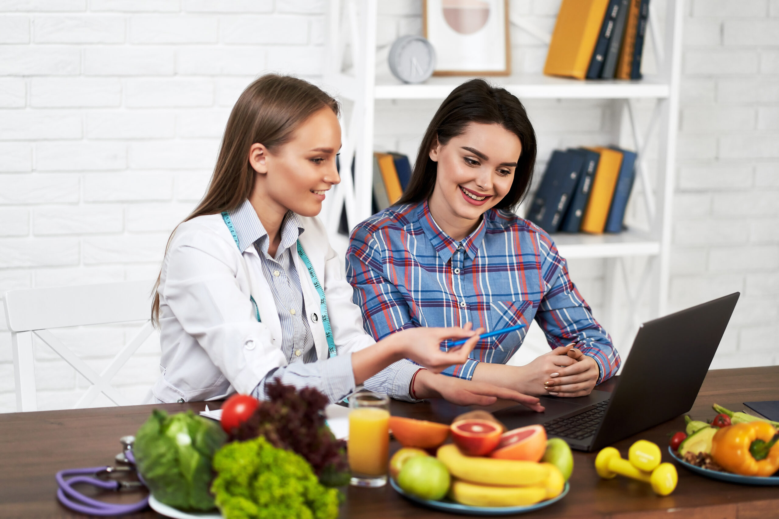 A smiling nutritionist advises a young patient woman on proper nutrition and dieting. The doctor shows in laptop a scheme of weight loss without a bad effect on health. 
Diet based on vegetables and fruits for girls.